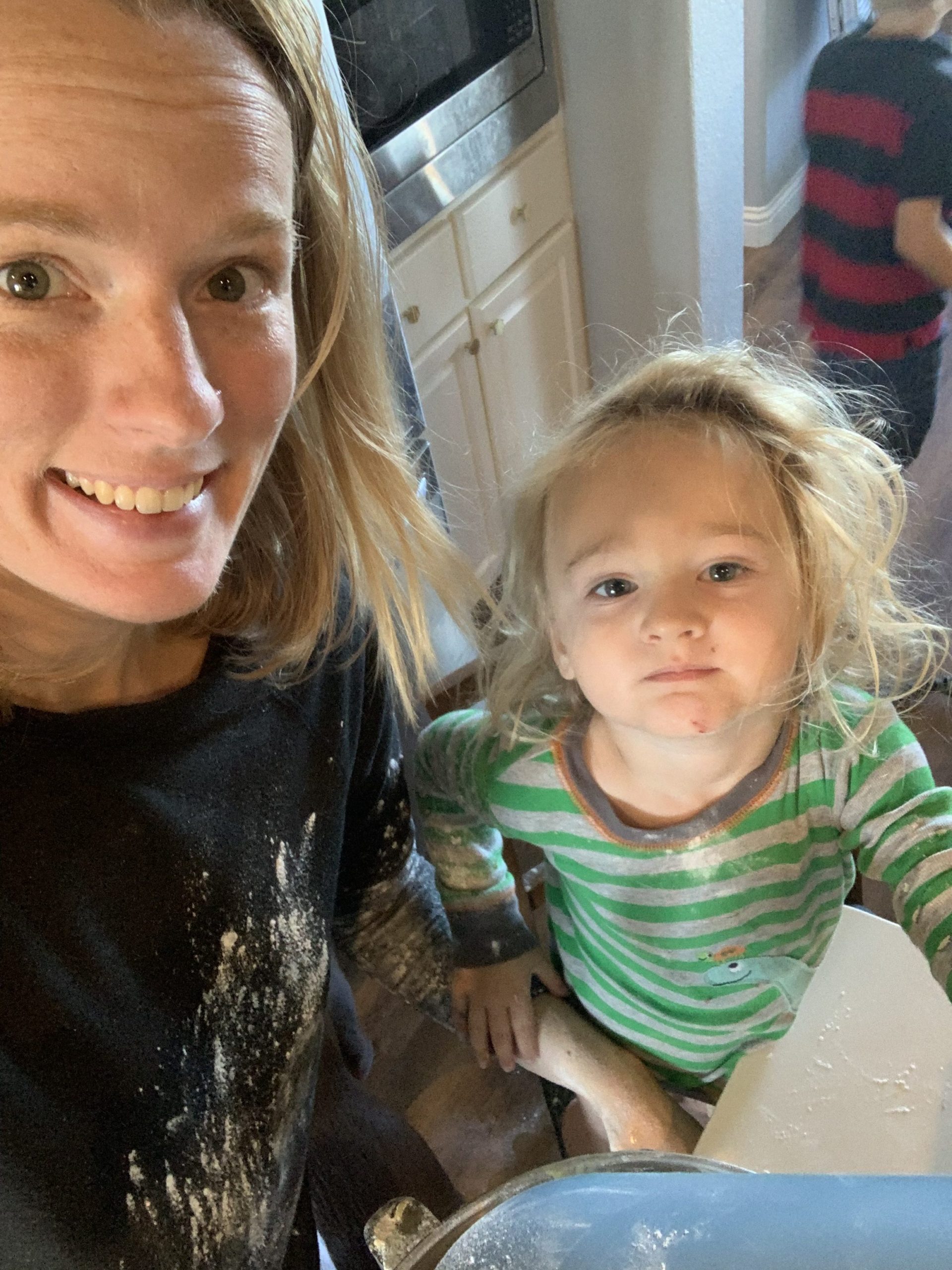 Mother in black shirt covered in flour with her toddler son standing next to her on a chair helping her in  the kitchen
